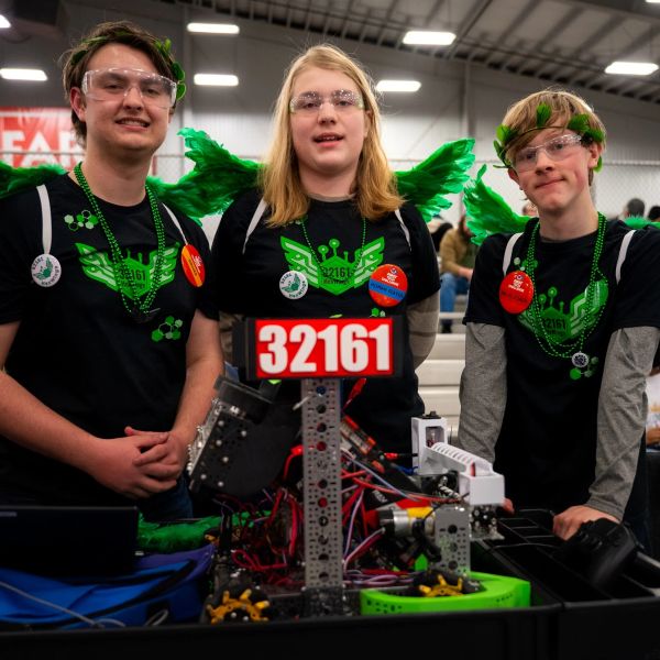 Three teens in matching shirts and green wings at a robotics event.