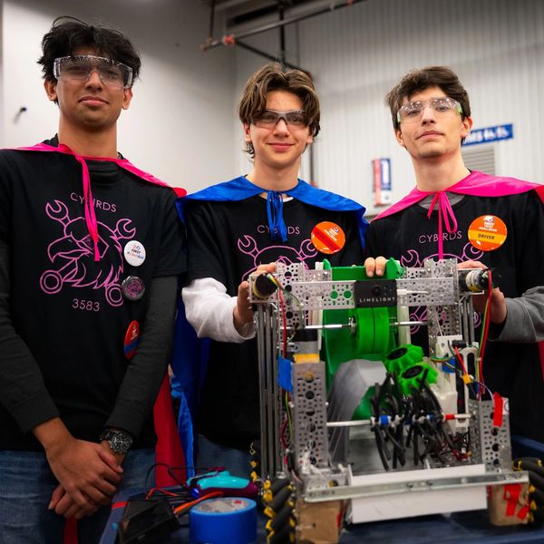 Three young men wearing safety glasses and capes with a robot.