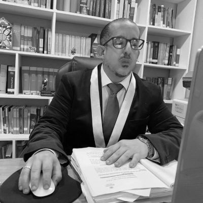 Man in suit and glasses working at a desk with bookshelves behind.