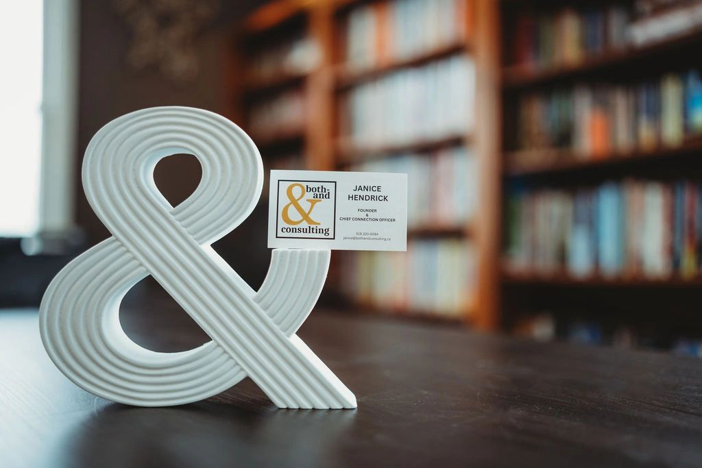 White ampersand holding a business card on a wooden table with blurred bookshelves.