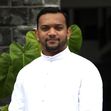 Man in white traditional attire standing outdoors with green leaves behind.