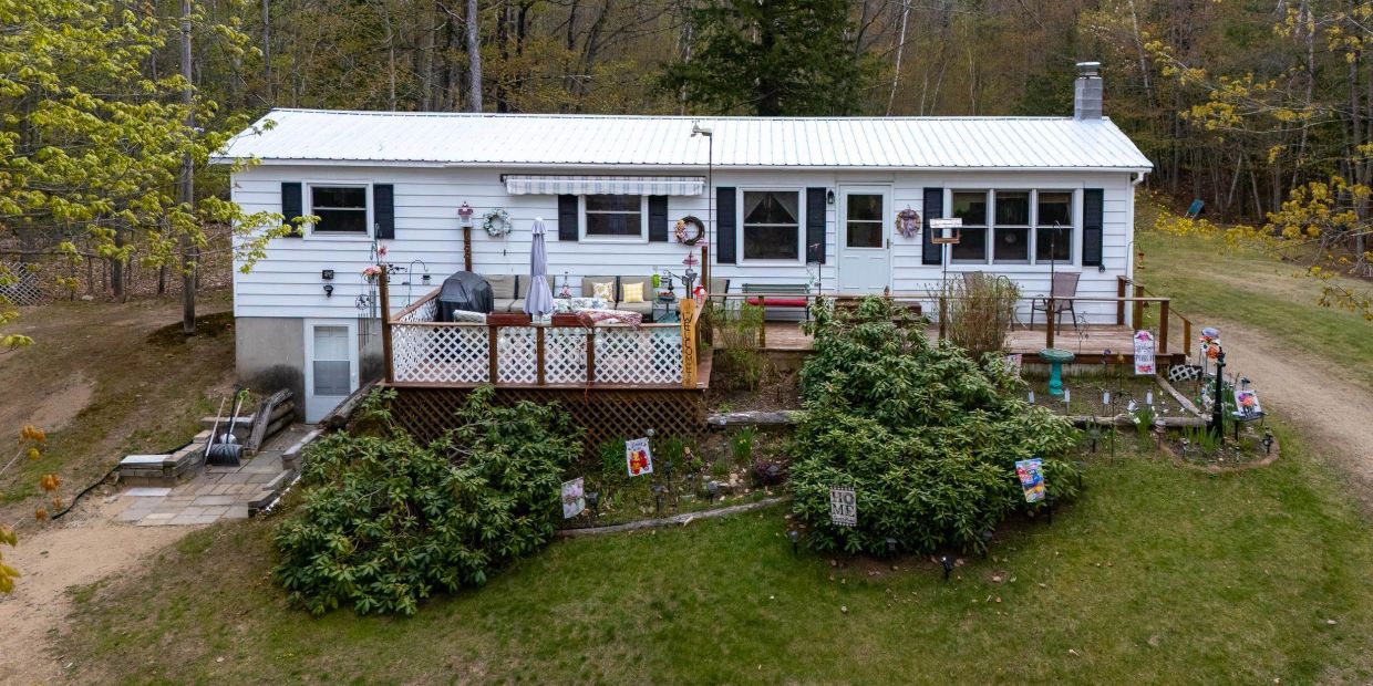 Cozy white house with a wooden deck and lush garden in a wooded area.