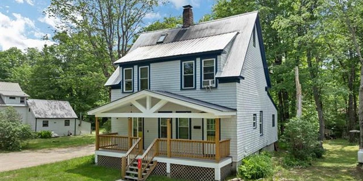 A white two-story house with a wooden porch surrounded by greenery.