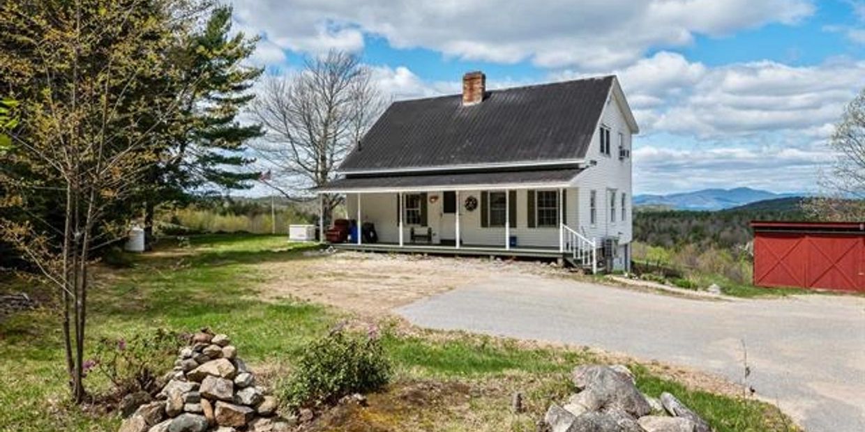 A white farmhouse with a black roof and a red shed in a rural setting.