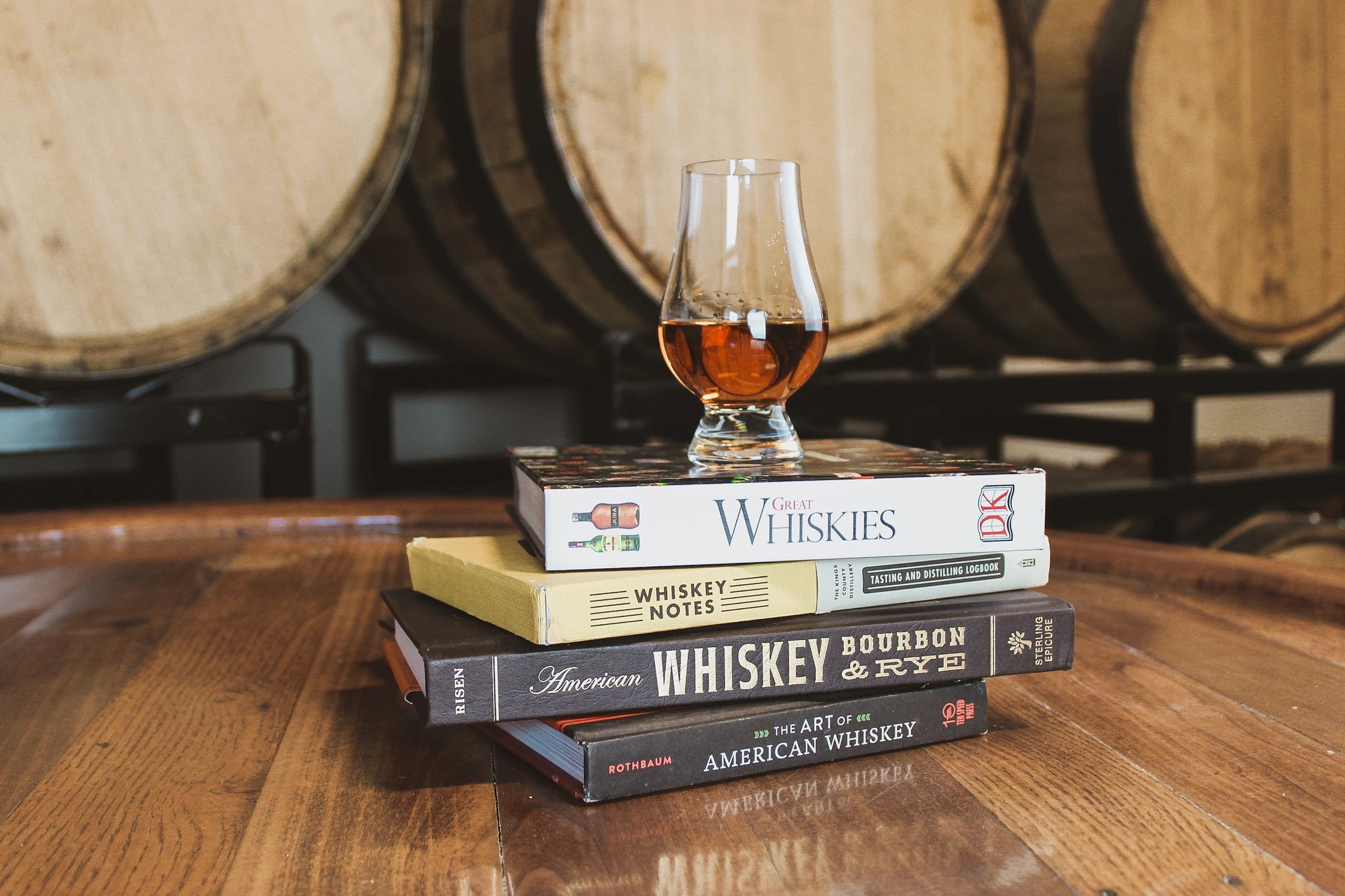 A glass of whiskey atop a stack of whiskey books on a wooden table.
