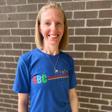 Smiling woman in blue ABC Pediatric Therapy t-shirt against a brick wall.