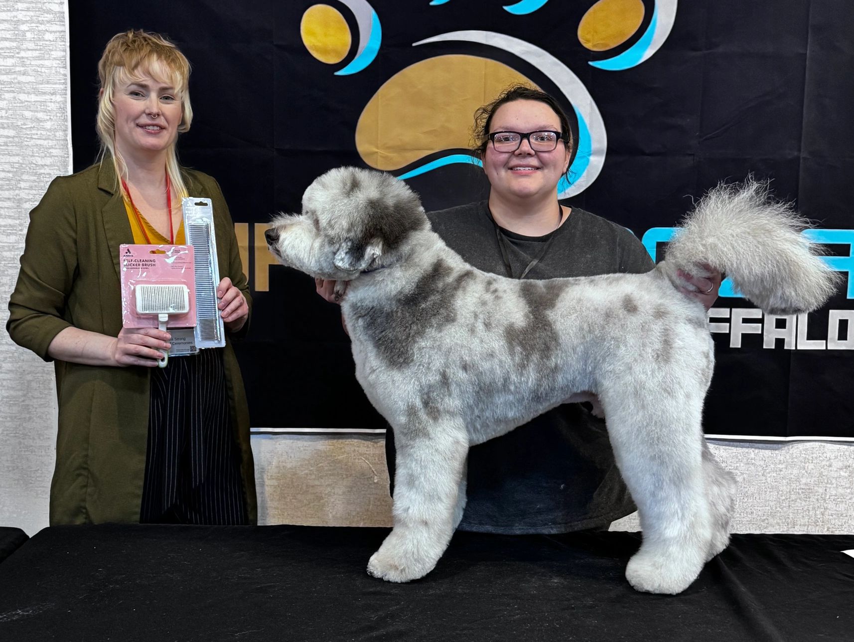 Two women proudly showcase a groomed dog and grooming tools at a pet grooming event.