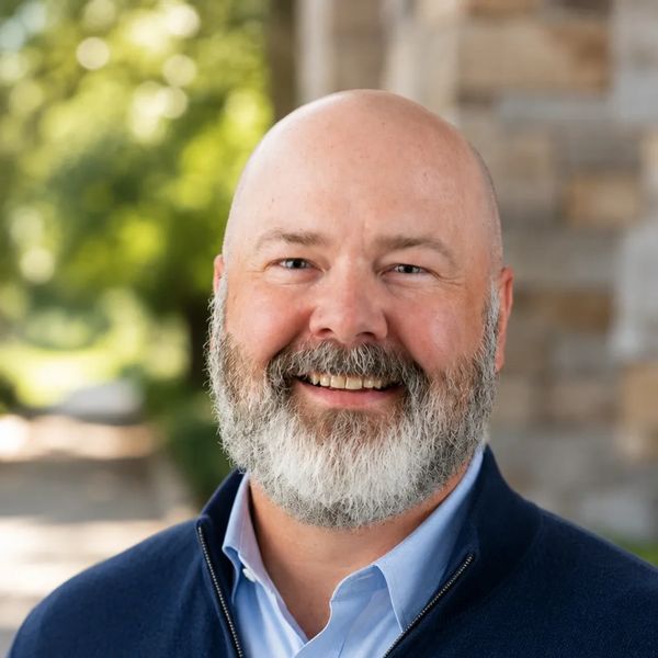 Smiling bald man with a gray beard outdoors in casual attire.