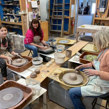 Three women happily working on pottery wheels in a ceramics studio.