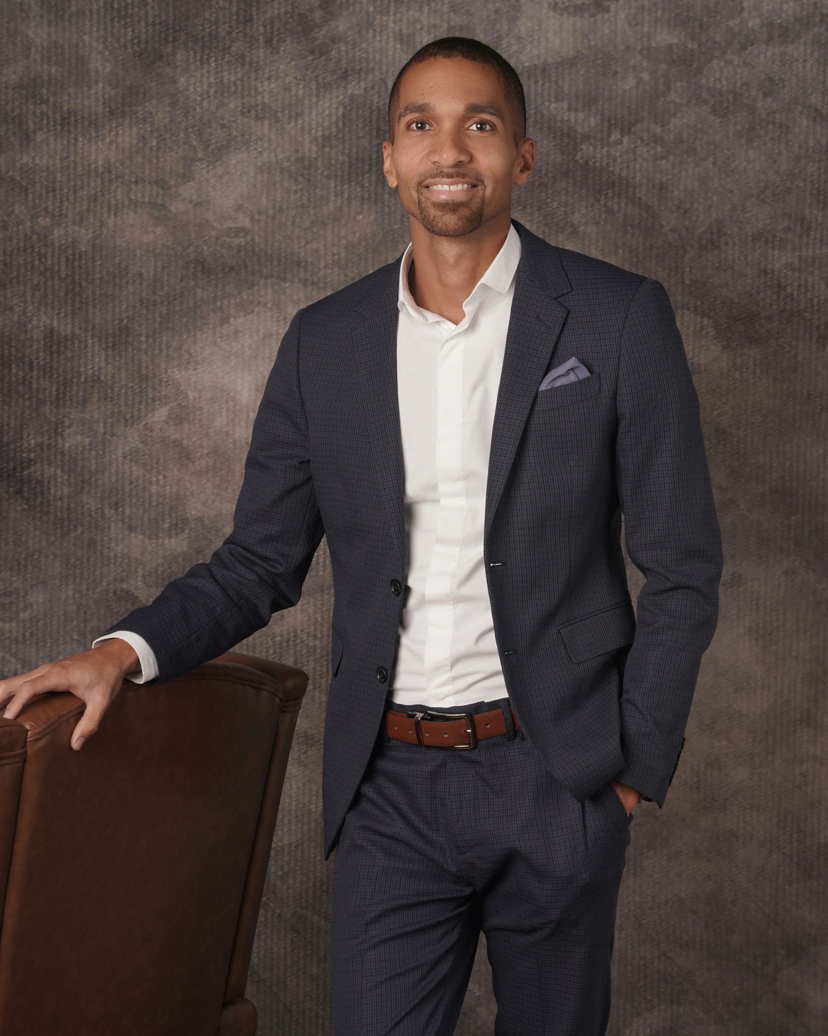 Young man in a tailored dark suit posing beside a leather chair.