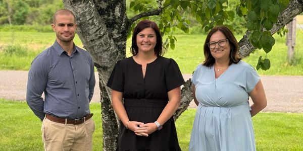 Three people standing outdoors in front of a tree, smiling.