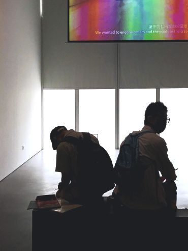 Two individuals sit back-to-back in a dimly lit gallery space with bright windows.