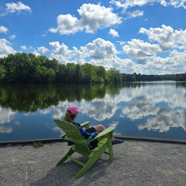 Person relaxing on a green chair by a reflective lake.