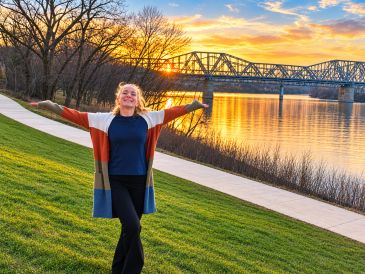 Woman enjoying sunset by the river with a bridge in the background.