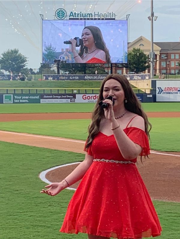 Woman in a red dress singing at a baseball field.