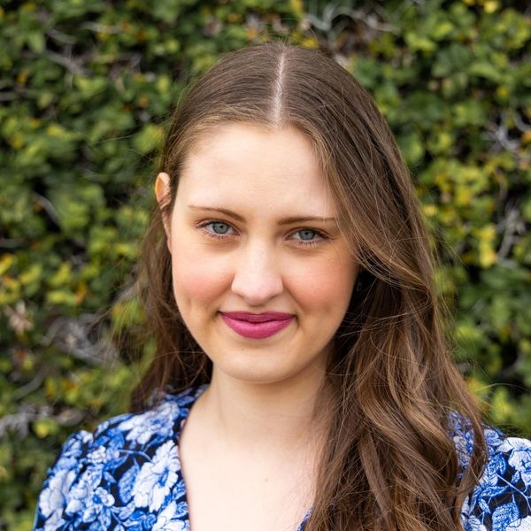 A headshot of Miriam Rogge wearing a floral blue-colored top with greenery in the background.