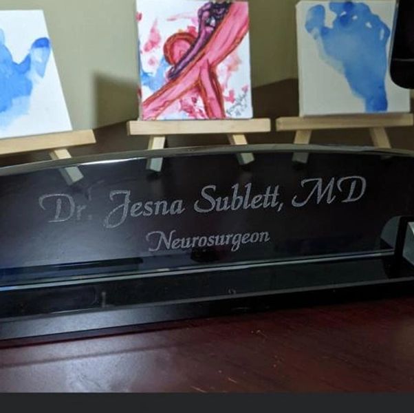 Desk nameplate for Dr. Jesna Sublett, MD, Neurosurgeon, with three small abstract paintings behind.