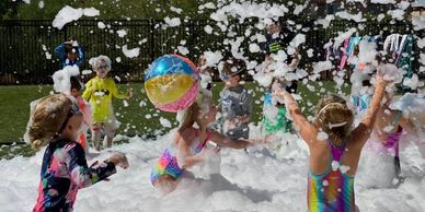 Children playing joyfully in foam with colorful beach balls outdoors.