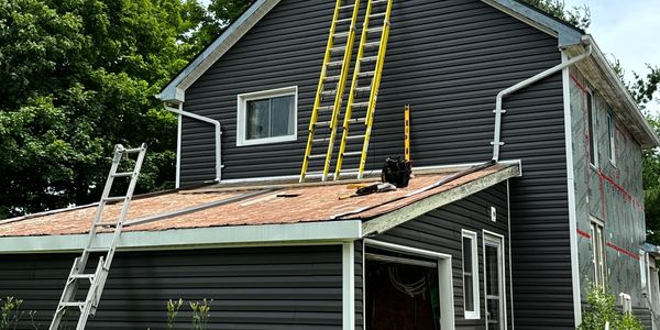 A black house exterior with ladders leaning against it and green trees in the background.