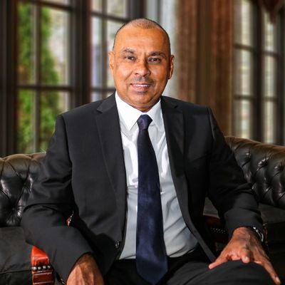 Man in a black suit sitting on a leather couch in a formal setting.