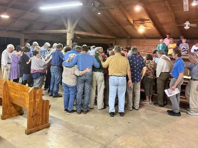 A group of people gathered closely in a wooden hall, some with arms around each other.