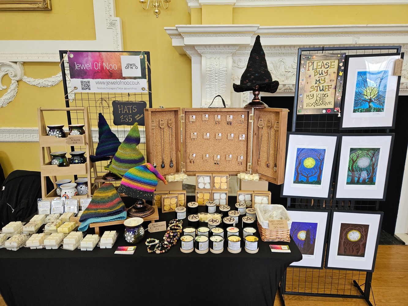 Craft fair stall with colorful handmade hats, jewelry, candles, and art prints.