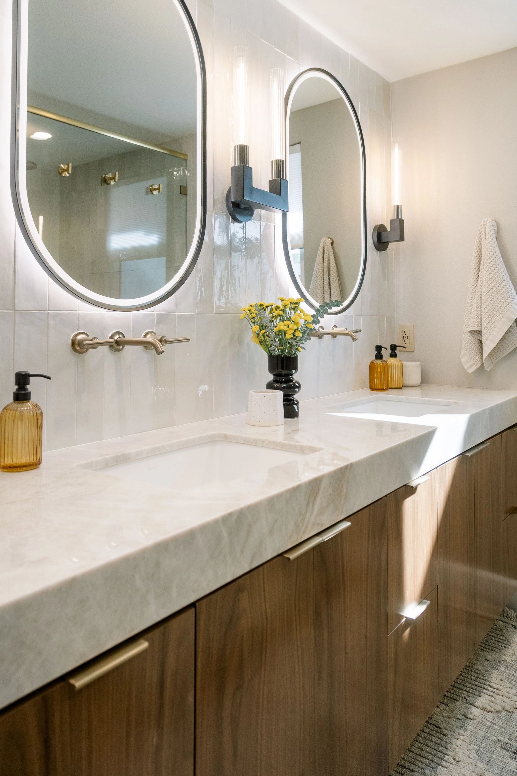 Double sink with bright mirrors, integrated lighting, white quartz countertop, and brown cabinetry.