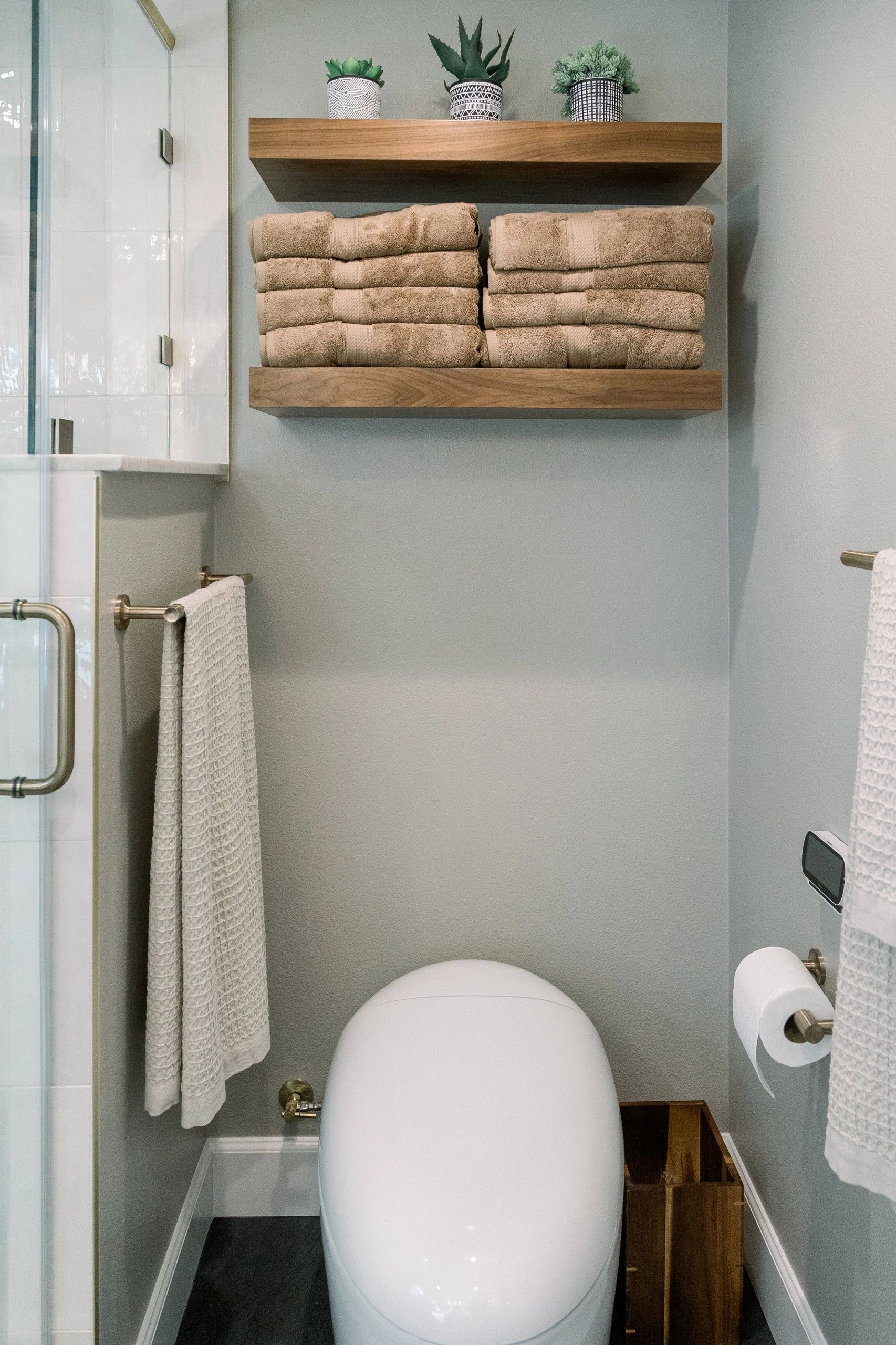 View of toilet area with grey walls, hanging shelf above holding folded brown towels and plants.