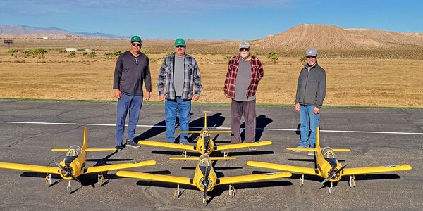 Four men stand behind four yellow model airplanes on a runway under a blue sky.