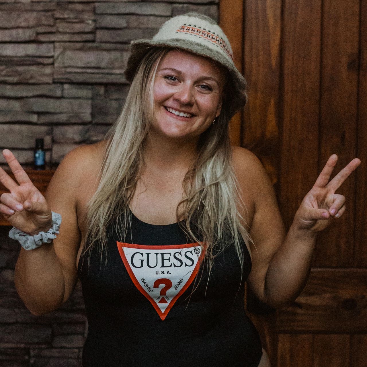 a woman wearing a black tank top and hat smiling and doing the peace sign with both her hands