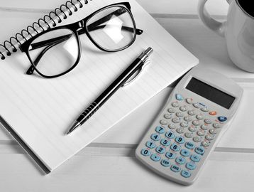 Black and white image of a notebook, pen, glasses, calculator, and coffee cup on a desk.