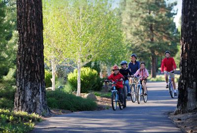 Family biking together on a sunny park trail surrounded by trees.