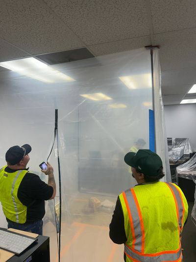 Two workers in safety vests inspect a plastic barrier in an indoor workspace.