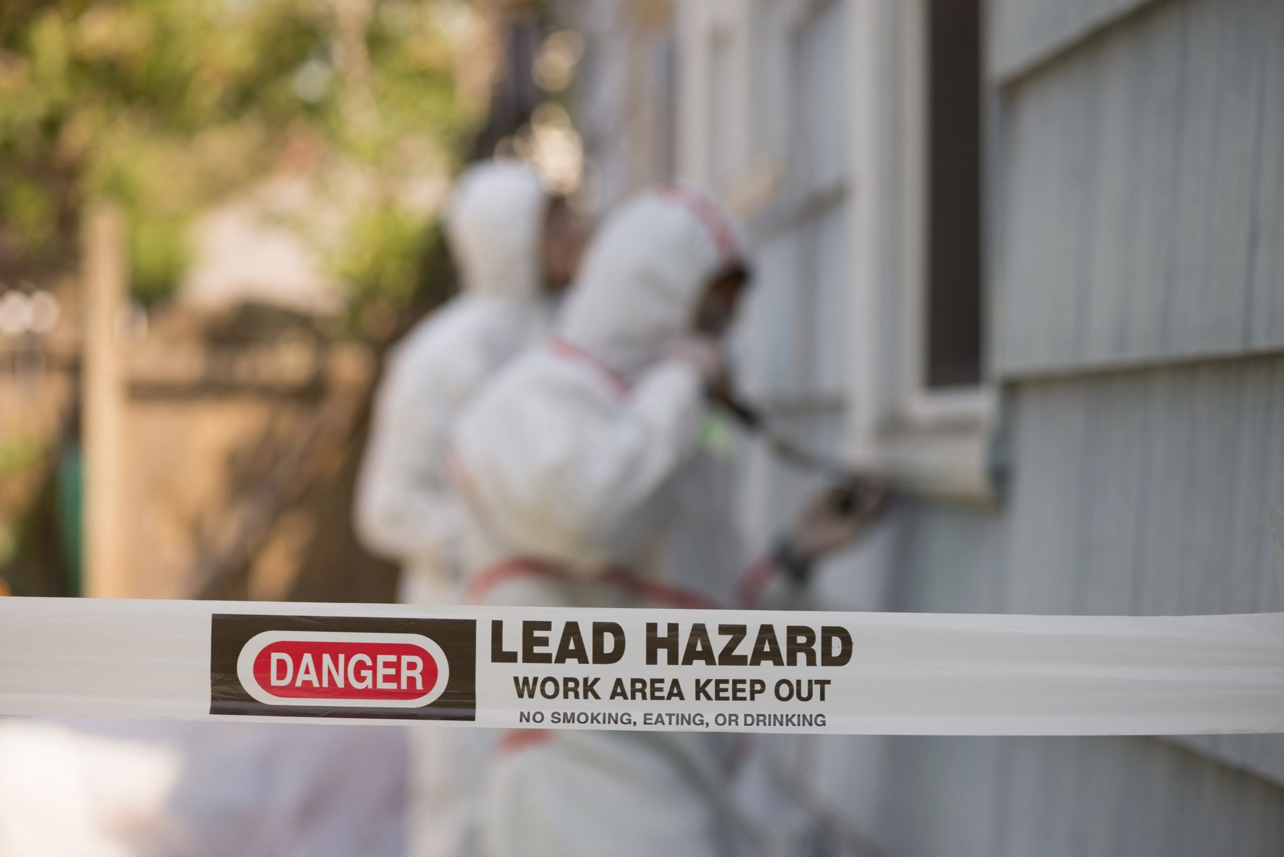Danger tape indicating a lead hazard work area with workers in protective suits.