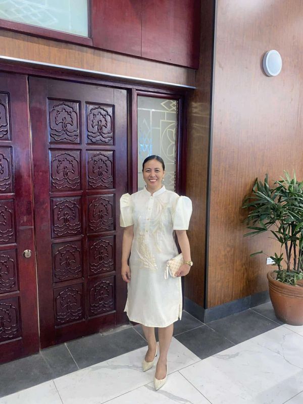 Woman in a white dress standing indoors near a wooden carved door and potted plant.