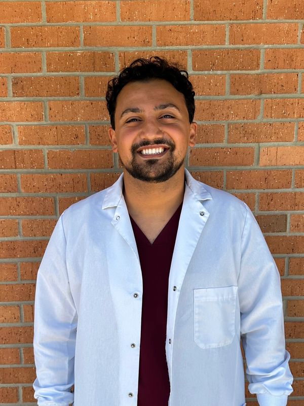 Smiling man in a white lab coat against a brick wall.