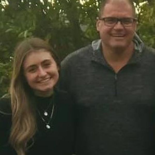 A smiling young woman and a man standing outdoors with trees in the background.