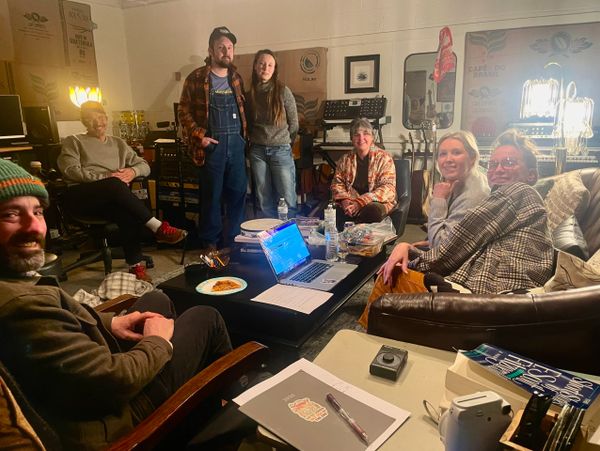 A group of seven people gathered in a cozy room around a table with a laptop and food.