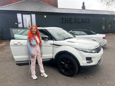 Woman with bright orange hair standing next to a white Range Rover.