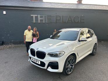A couple posing beside a white BMW outside a building named The Place.