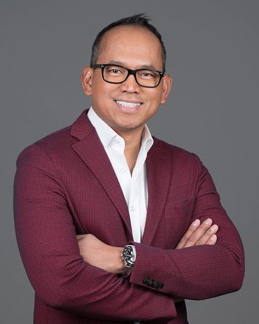Smiling man in a burgundy blazer and glasses poses confidently with arms crossed.