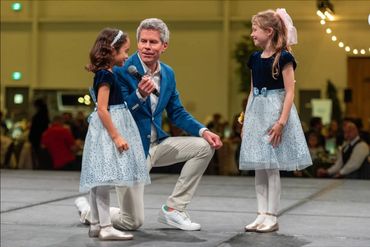 Man kneels, speaking to two young girls in matching dresses on stage.