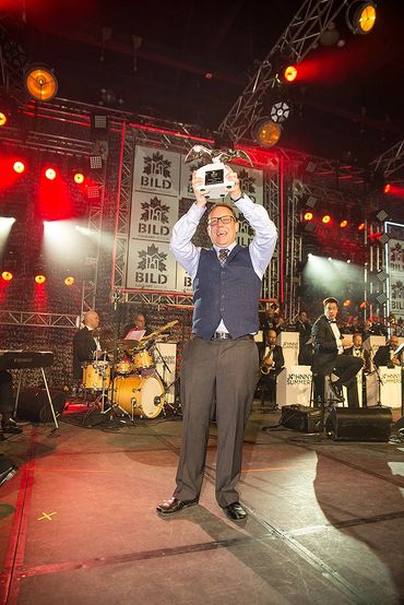 Man in formal attire holding up a trophy on stage with band in background.