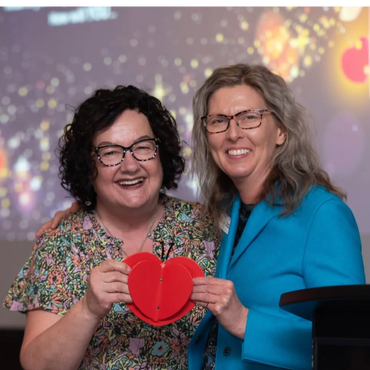 Two women smiling and holding a red heart-shaped object.
