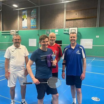 Group of four men posing with badminton equipment indoors.