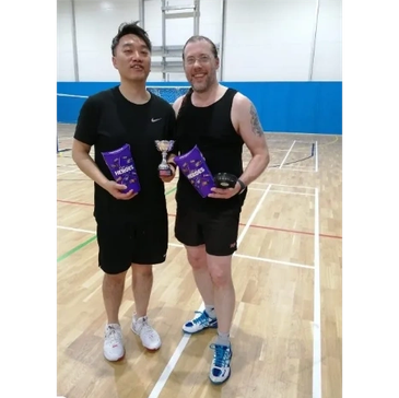 Two men holding trophies and chocolate on an indoor court.