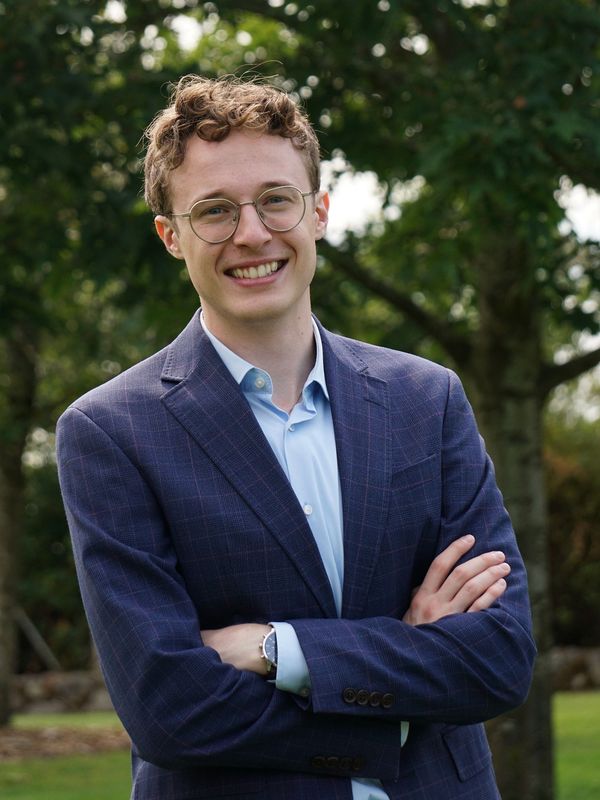 Young man with glasses smiling outdoors in a blue suit.