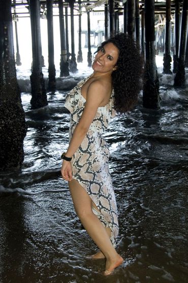 Woman smiling in a patterned dress standing barefoot in shallow water under a pier.