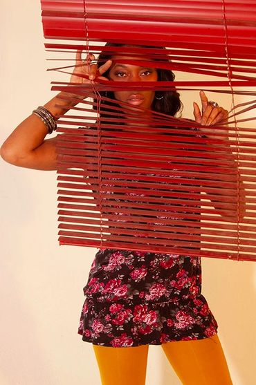 A woman peeks through red window blinds, wearing a floral dress and yellow tights.