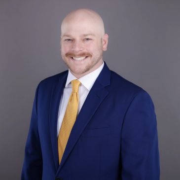 Smiling bald man in blue suit and yellow tie against gray background.